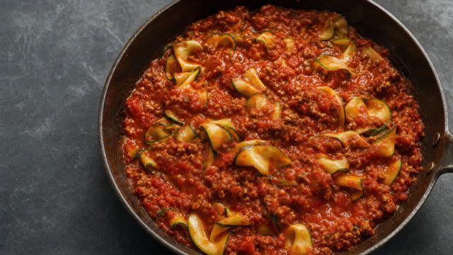 Pfanne mit Zucchinibändern in roter Bolognese auf dunklem Untergrund, seitlich beleuchtet.