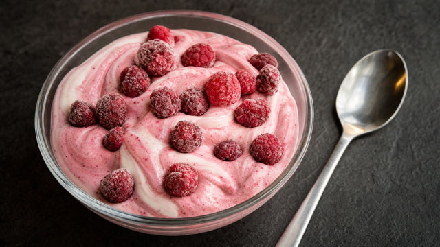 Glasschale mit rosa marmorierter Protein-Mousse und gefrorenen Himbeeren, Löffel daneben, dunkler Untergrund.