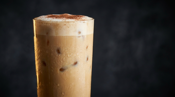 Cremiger X-FORMING Protein-Eiskaffee im hohen Glas mit Eiswürfeln, Milchschaum und Kakao auf dunklem Hintergrund mit Gold-Akzent
