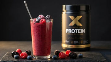 Hohes Glas mit cremigem Beeren-Protein-Smoothie, obenauf frische Himbeeren und Brombeeren, daneben die X-PROTEIN Natur Dose auf dunklem Schiefer