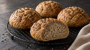 Vier goldbraune Brötchen mit Sesam und Kümmel auf einem schwarzen Kuchengitter, eines aufgeschnitten mit feinporiger Krume, dunkler Schieferuntergrund.