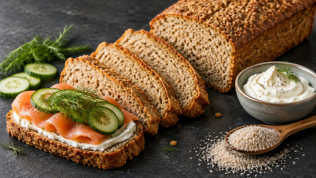 Kastenbrot ohne Mehl, angeschnitten, vier Scheiben fächerförmig davor, eine Scheibe belegt mit Frischkäse, Räucherlachs, Gurke und Dill, daneben Frischkäse und ein Löffel Flohsamenschalen auf dunklem Schiefer.