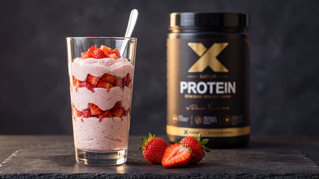 Hohes Glas mit geschichtetem Erdbeer-Protein-Dessert, daneben die X-PROTEIN Natur Dose und frische Erdbeeren auf dunklem Schiefer