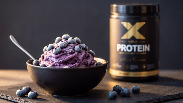 Dunkle Schale mit cremiger Heidelbeer-Protein-Frozen-Bowl und gefrorenen Heidelbeeren, daneben die X-PROTEIN Natur Dose auf dunklem Schiefer