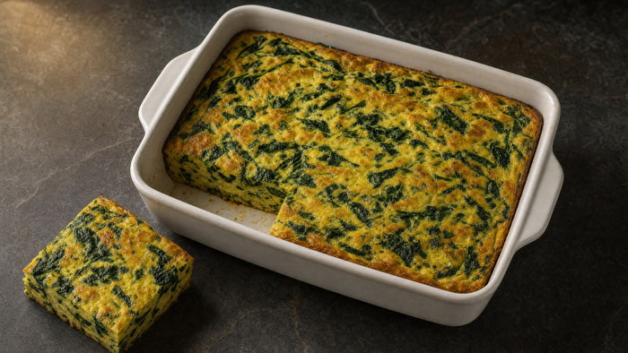 Goldbraune Spinat-Frittata in einer weißen Auflaufform, ein Stück herausgeschnitten und daneben gelegt, dunkler Steinuntergrund.