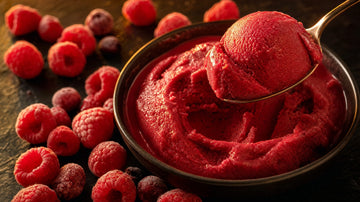 Dunkle Schale mit cremigem Himbeer-Protein-Eis, ein Löffel hebt eine Kugel heraus, ringsum gefrorene Himbeeren im warmen Gegenlicht