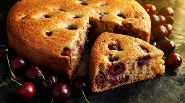 Angeschnittener Protein-Kirschkuchen auf dunklem Schiefer, ein Stück herausgezogen mit sichtbaren Sauerkirschen, warmes Licht von rechts