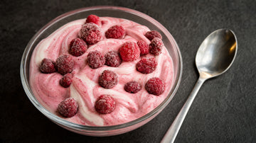 Glasschale mit rosa marmorierter Protein-Mousse und gefrorenen Himbeeren, Löffel daneben, dunkler Untergrund.