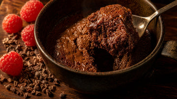Dunkle Tasse mit warmem Schoko-Protein-Tassenkuchen, ein Löffel hebt ein Stück heraus, daneben Himbeeren und Kakao-Nibs im warmen Abendlicht