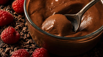 Löffel taucht in cremige dunkle Schoko-Protein-Creme im Glas, Himbeeren und Kakao-Nibs daneben
