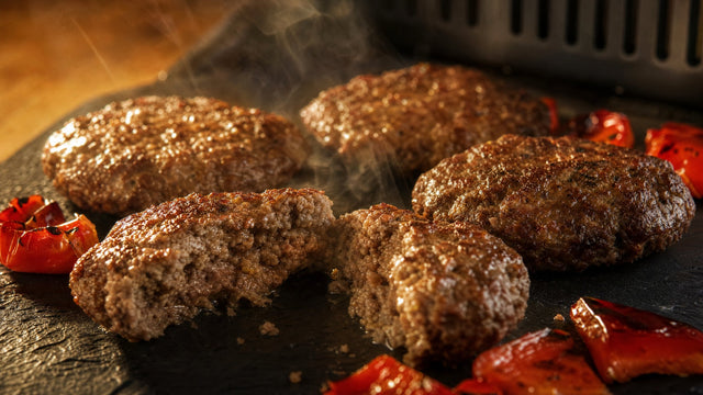Vier flache Tatar-Frikadellen auf dunklem Schiefer, die vordere aufgebrochen mit saftigem Inneren, geröstete rote Paprika daneben