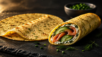 Zwei Wraps ohne Mehl auf dunklem Schiefer, einer flach liegend mit gesprenkelter goldgelber Oberfläche, einer gerollt und angeschnitten mit Räucherlachs, Gurke, Kräutern und Frischkäse.